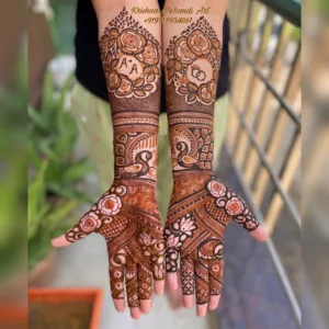 Intricate bridal henna design by Krishna Mehandi Art, a bridal mehndi artist in Kasauli. The image displays customized full-hand and forearm patterns including the couple's initials, wedding rings in rose wreaths, traditional peacock motifs on the wrists, and lotus designs on the palms, ideal for modern Indian wedding bridal henna inspiration.